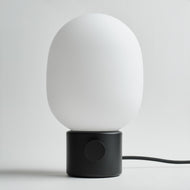 The Audo Opal Table Lamp features a smooth white oval shade, round black base, and a visible black cord extending to the right, all set against a plain light background.