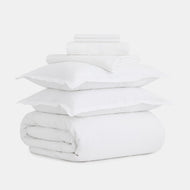 The Linen Foundational Bedding Set, featuring two pillows, a folded comforter, and sheets, is neatly stacked and displayed on a plain white background.