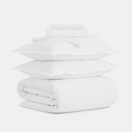 The Cotton Percale Foundational Bedding Set, featuring two pillows, a folded duvet, and sheets, is neatly stacked against a plain white background.