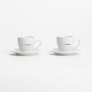 The Café Thuma Espresso Set features two white ceramic cups with handles on matching saucers, each branded with "CAFÉ THUMA" on the side, all set against a plain white background.