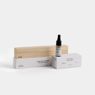A dropper bottle labeled From Soil to Form Oil Diffuser Charcoal by Frama sits on white boxes with minimalist text. A rectangular wooden block stands upright behind the boxes, all set against a plain white background.