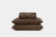 The Cotton Ribbed Coverlet Set, in dark brown with a textured ribbed pattern, includes a neatly folded blanket and two matching pillows set against a plain light gray background.