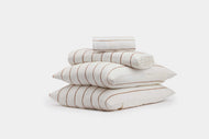The Linen Stripe Sheet Set by CULTIVER is displayed neatly stacked with two pillows, a folded flat sheet, and a rolled sheet, all in white with thin brown horizontal stripes against a plain white background.