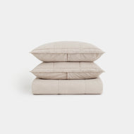 Two beige pillows are stacked on a folded Linen Box Quilt Set, all arranged against a plain white background.