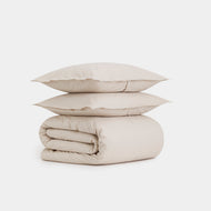 The Cotton Percale Duvet Cover Set is displayed with two cream-colored pillows stacked atop a neatly folded comforter, all set against a plain white background.
