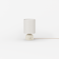 The Ceramic Table Lamp, featuring a cylindrical white fabric shade, round white base, and small button, sits on a white surface with its cord extending to the right.