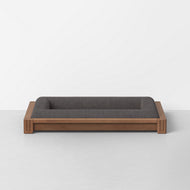 The Classic Dog Bed features a wooden frame with a dark gray cushioned mattress, shown here on a light gray floor against a plain white wall.