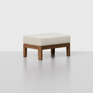 The Classic Ottoman features a modern rectangular design with a light beige cushioned top and a simple wooden frame, displayed on a smooth gray surface against a plain light background.