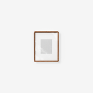 The Cove Frame is a simple brown wooden photo frame with a white mat and empty center, hanging on a plain white wall. Soft shadows appear on the mat from light coming from the left.