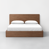 The Essential Bed + Headboard, featuring a modern wooden design and high headboard, is neatly dressed in white sheets and two pillows, set against a plain white wall and light gray floor.