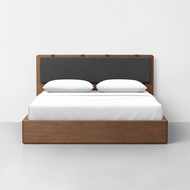 The Essential Bed + Headboard + Cushion features a modern wooden frame, dark upholstered headboard, white bedding, and two white pillows, all set against a plain white wall in a minimalistic room.