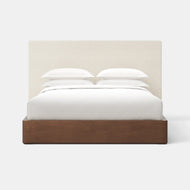 The Essential Bed + Upholstered Headboard features a tall, light-toned fabric headboard and a wooden platform base, elegantly styled with white sheets and pillows, set against a plain white background.