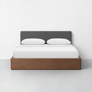 The CL Test - Essential Bed features a dark gray upholstered headboard, white bedding with two pillows, and a wooden platform base, set against a plain white wall and light gray floor.