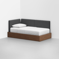 The Essential Daybed features a wooden base, gray upholstered backrest, and white bedding with a pillow, perfectly positioned in the corner of a minimalist room with white walls and a light gray floor.