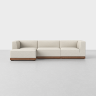 The Essential Chaise Sectional, a modern light beige sofa with clean lines and wooden base, sits on a smooth light gray floor against a plain white background.