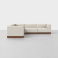 The Essential Corner Sectional features light beige upholstery and a wooden base, showcased in a minimalist room with a light gray floor and white walls.