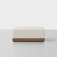 The Essential Ottoman features a cream-colored upholstered cushion on a modern rectangular wooden base, shown against a plain white background.