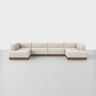 The Essential U Sectional is a large, modern sofa with light beige upholstery and a wooden base, arranged in an L-shape on a smooth light gray floor against a plain white background.
