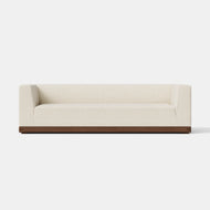 The Essential Sofa, 96″ is a modern, cream-colored sofa with a boxy design and low, straight armrests, featuring a dark wooden base and set against a plain white background.