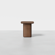 The Pedestal Side Table features a minimalist design with a square wooden top and a single cylindrical base, displayed against a plain white background.