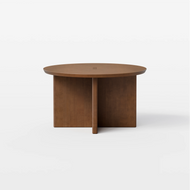 The Round Pillar Coffee Table is a round wooden coffee table with a simple, modern design, featuring a thick tabletop and intersecting panel legs, set against a plain white background.