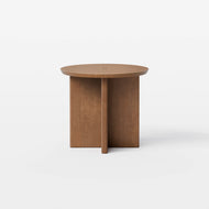 The Pillar Side Table is a modern round wooden side table featuring a smooth top and a distinctive intersecting panel base, displayed against a plain white background.