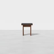 A Pillar Stool with a dark rectangular wooden top and simple legs sits in the center of an empty, light-colored room with a minimal background.
