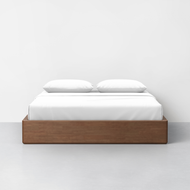 The Essential Storage Bed Frame, featuring a sleek wooden design and crisp white bedding, is positioned against a plain white wall with two pillows neatly arranged on top.