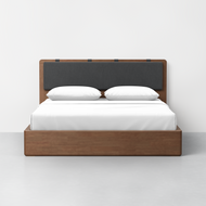 The Essential Storage Bed features a wooden frame paired with a dark gray upholstered headboard and cushion, topped with crisp white bedding and two pillows, all set against a plain white wall and a light gray floor.