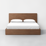 The Essential Storage Bed + Headboard, featuring a tall wooden headboard, is neatly made with white bedding and two white pillows, and is positioned on a light gray floor against a plain white wall.