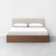 The Essential Storage Bed + Pillowboard features a contemporary wooden base, a light upholstered pillowboard headboard, crisp white bedding, and two pillows, all set against a plain white wall on a light-toned floor.