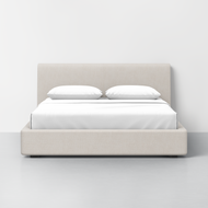 The Signature Bed features a light beige upholstered frame and headboard, made up with white sheets and two pillows, set against a plain white wall on a light gray floor.