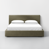 The Thuma x Buchanan Studio - Signature Bed features an olive green upholstered frame and headboard, neatly dressed in white bedding with two pillows, set against a plain white wall in a minimalistic room.