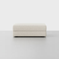 The Signature Storage Ottoman is a rectangular, light beige upholstered piece with a minimal, modern design and hidden legs, shown on a light grey floor against a plain white background.