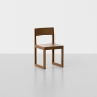 The Studio Chair is a modern wooden chair featuring a square seat, straight backrest, and rectangular legs, set on a smooth light gray floor against a plain white background.