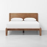 The Classic Bed features a wooden frame with a simple headboard, topped with a white mattress and two white pillows, placed against a plain white wall and floor.