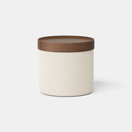 The Upholstered Ottoman + Tray features a round, cream-colored fabric body paired with a smooth, flat brown wooden lid, displayed against a plain white background.