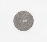 A round, gray Felt Coaster featuring the word THUMA in white, displayed on a plain white background.